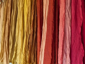A vibrant display of colorful fabrics hanging vertically, ranging in shades from yellow to deep red. The fabrics appear to have a textured surface with slight wrinkles, suggesting natural fiber material.
