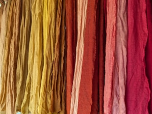 A vibrant display of colorful fabrics hanging vertically, ranging in shades from yellow to deep red. The fabrics appear to have a textured surface with slight wrinkles, suggesting natural fiber material.
