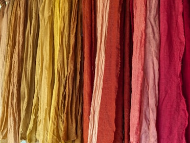 A vibrant display of colorful fabrics hanging vertically, ranging in shades from yellow to deep red. The fabrics appear to have a textured surface with slight wrinkles, suggesting natural fiber material.