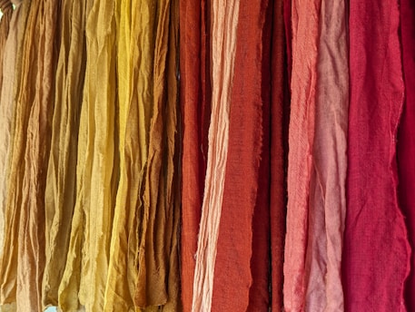 A vibrant display of colorful fabrics hanging vertically, ranging in shades from yellow to deep red. The fabrics appear to have a textured surface with slight wrinkles, suggesting natural fiber material.