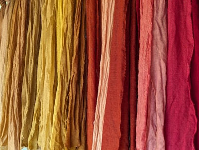 A vibrant display of colorful fabrics hanging vertically, ranging in shades from yellow to deep red. The fabrics appear to have a textured surface with slight wrinkles, suggesting natural fiber material.