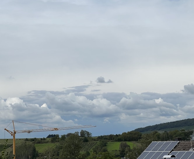 A rural landscape with overcast skies and scattered clouds. A large construction crane stands prominently on the left, with solar panels visible on the rooftops to the right. The background features rolling hills and dense green foliage.
