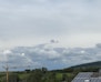 A rural landscape with overcast skies and scattered clouds. A large construction crane stands prominently on the left, with solar panels visible on the rooftops to the right. The background features rolling hills and dense green foliage.
