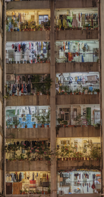 A multi-story residential building with several balconies filled with various items. Each balcony is decorated with potted plants and clothes hanging to dry. The building's facade appears weathered, and there are visible metal railings. The lighting inside suggests evening or nighttime, with warm and cool lights illuminating different units, adding a cozy and lived-in atmosphere.
