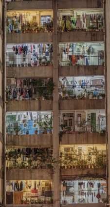 A multi-story residential building with several balconies filled with various items. Each balcony is decorated with potted plants and clothes hanging to dry. The building's facade appears weathered, and there are visible metal railings. The lighting inside suggests evening or nighttime, with warm and cool lights illuminating different units, adding a cozy and lived-in atmosphere.