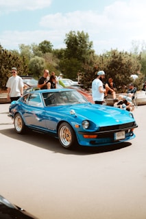 A blue classic car with golden rims is parked outdoors, with a small gathering of people around it. The background features trees and other cars, indicating an outdoor car show or gathering. Several individuals are standing, sitting or leaning against barriers, casually enjoying the event under a partly cloudy sky.