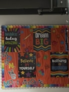 A bulletin board with colorful motivational posters and decorations. The posters include messages such as 'Make today amazing,' 'Dream big,' 'Believe in yourself,' and 'Nothing is impossible.' The board is adorned with illustrations of stars, hands, and vibrant text, all set against an orange background.