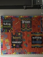 A bulletin board with colorful motivational posters and decorations. The posters include messages such as 'Make today amazing,' 'Dream big,' 'Believe in yourself,' and 'Nothing is impossible.' The board is adorned with illustrations of stars, hands, and vibrant text, all set against an orange background.