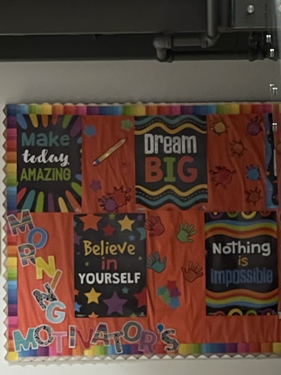 A bulletin board with colorful motivational posters and decorations. The posters include messages such as 'Make today amazing,' 'Dream big,' 'Believe in yourself,' and 'Nothing is impossible.' The board is adorned with illustrations of stars, hands, and vibrant text, all set against an orange background.