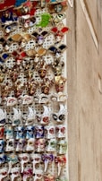 A vertical display of intricately decorated Venetian masks featuring a variety of colors including red, gold, white, blue, and green. Each mask has unique embellishments, such as glitter, feathers, and ornate designs. The masks are arranged in rows and create a vibrant and festive visual.