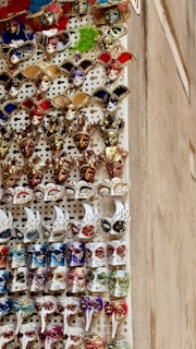 A vertical display of intricately decorated Venetian masks featuring a variety of colors including red, gold, white, blue, and green. Each mask has unique embellishments, such as glitter, feathers, and ornate designs. The masks are arranged in rows and create a vibrant and festive visual.