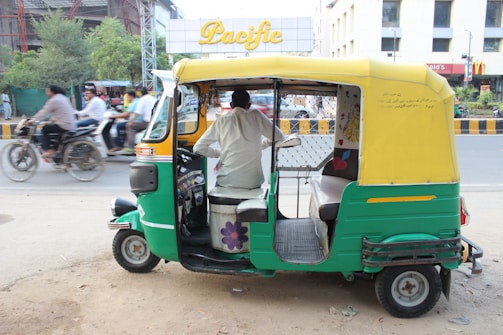 A green and yellow auto-rickshaw is parked on the side of the road with the driver seated inside. In the background, there is a busy street scene with several motorcycles and bicycles in motion. Buildings with signage are visible, including a sign that reads 'Pacific'.