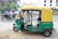 A green and yellow auto-rickshaw is parked on the side of the road with the driver seated inside. In the background, there is a busy street scene with several motorcycles and bicycles in motion. Buildings with signage are visible, including a sign that reads 'Pacific'.