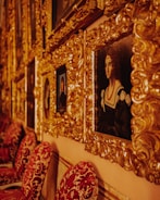 A series of ornate gold-framed paintings are displayed on a wall. The frames are intricately designed with a baroque style. A prominent portrait of a woman in historical attire is visible, featuring a black dress with white details and a red necklace. The wall behind the frames is a warm color, complementing the luxurious texture and color of the frames. Below the paintings, red velvet chairs with intricate patterns are aligned against the wall.