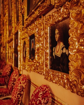 A series of ornate gold-framed paintings are displayed on a wall. The frames are intricately designed with a baroque style. A prominent portrait of a woman in historical attire is visible, featuring a black dress with white details and a red necklace. The wall behind the frames is a warm color, complementing the luxurious texture and color of the frames. Below the paintings, red velvet chairs with intricate patterns are aligned against the wall.
