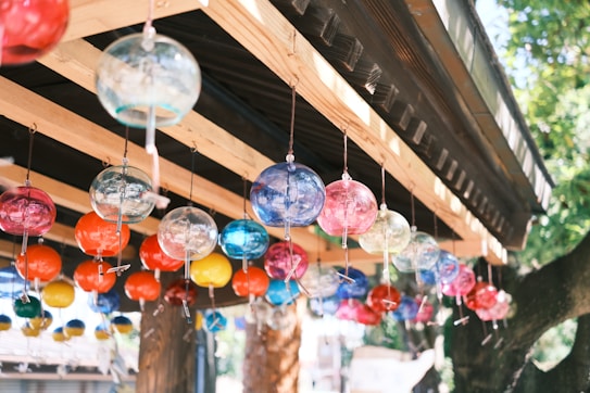 A variety of colorful glass wind chimes hang from the wooden beams of an outdoor wooden structure. The structure is shaded by a roof, and there are trees and other elements of nature around it.