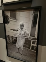 An elderly woman is using a hula hoop in a yard. She is wearing a patterned dress and appears focused on the activity. The background shows a house with a white exterior and a brick foundation. The photo is in black and white, adding a nostalgic feel.