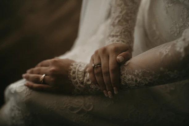 Close-up of delicate wedding details: lace dress sleeve and intertwined hands against a soft natural backdrop.