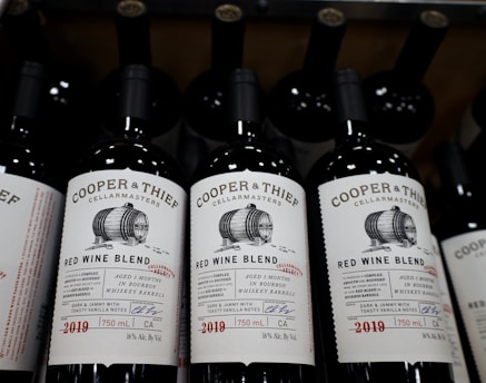 Several bottles of Cooper & Thief Red Wine Blend are displayed, featuring a label with an illustration of a barrel. The label highlights that the wine is aged in bourbon whiskey barrels and contains 16% alcohol by volume. The year 2019 is noted along with a signature and details about the wine.