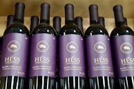 Several bottles of red wine are displayed in a row. The labels on the bottles are purple with gold lettering, indicating they are part of The Hess Collection, specifically the Allomi Napa Valley Cabernet Sauvignon from 2019.