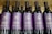 Several bottles of red wine are displayed in a row. The labels on the bottles are purple with gold lettering, indicating they are part of The Hess Collection, specifically the Allomi Napa Valley Cabernet Sauvignon from 2019.
