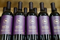 Several bottles of red wine are displayed in a row. The labels on the bottles are purple with gold lettering, indicating they are part of The Hess Collection, specifically the Allomi Napa Valley Cabernet Sauvignon from 2019.