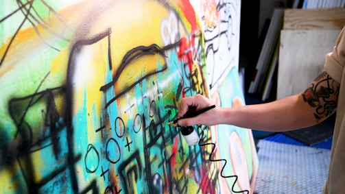 A person with a tattooed arm is using an airbrush to paint on a colorful canvas filled with abstract shapes and symbols. The artwork is vibrant and dynamic, featuring a mix of curves, lines, and various geometric forms.
