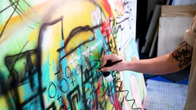 A person with a tattooed arm is using an airbrush to paint on a colorful canvas filled with abstract shapes and symbols. The artwork is vibrant and dynamic, featuring a mix of curves, lines, and various geometric forms.