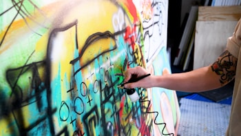 A person with a tattooed arm is using an airbrush to paint on a colorful canvas filled with abstract shapes and symbols. The artwork is vibrant and dynamic, featuring a mix of curves, lines, and various geometric forms.