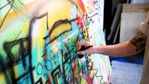 A person with a tattooed arm is using an airbrush to paint on a colorful canvas filled with abstract shapes and symbols. The artwork is vibrant and dynamic, featuring a mix of curves, lines, and various geometric forms.