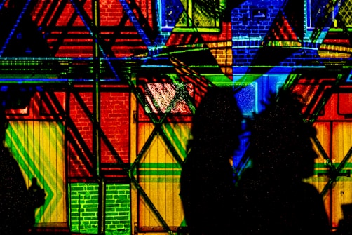 Abstract geometric patterns are projected onto a wall, creating a vibrant and colorful backdrop. The wall features intersecting lines and shapes in bold colors, including red, blue, yellow, and green. Silhouettes of two people stand in the foreground, creating an interesting contrast against the bright background.