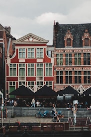 A picturesque scene featuring classic European architecture with intricate brickwork and large windows. People are sitting and walking near a waterside cafe, which has tables under black umbrellas. There are also a few bicycles visible on the pavement.