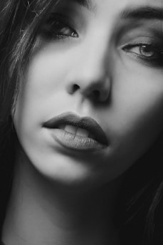 The image is a close-up black and white portrait of a woman's face, highlighting her eyes, lips, and skin texture. Her eyes have a reflective gaze with subtle makeup accentuating her eyelashes and eyebrows. The lighting creates shadows that contour her facial features, emphasizing a serene and introspective expression.