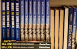 A neatly arranged bookshelf displaying a series of books with blue and yellow covers. The titles include 'Made in Sweden,' 'Universum och jag,' and 'Pappas Pojke.' A stack of smaller books labeled 'Stå Upp! Boken om Stand-up Comedy' with a dark cover is visible at the bottom.