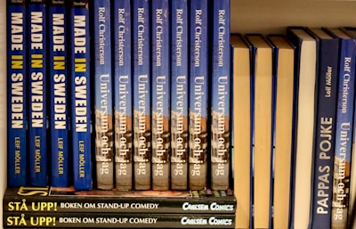 A neatly arranged bookshelf displaying a series of books with blue and yellow covers. The titles include 'Made in Sweden,' 'Universum och jag,' and 'Pappas Pojke.' A stack of smaller books labeled 'Stå Upp! Boken om Stand-up Comedy' with a dark cover is visible at the bottom.