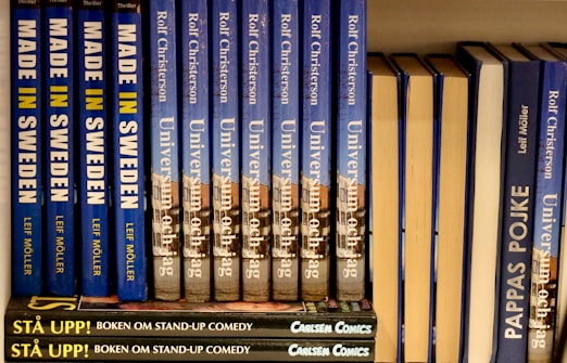 A neatly arranged bookshelf displaying a series of books with blue and yellow covers. The titles include 'Made in Sweden,' 'Universum och jag,' and 'Pappas Pojke.' A stack of smaller books labeled 'Stå Upp! Boken om Stand-up Comedy' with a dark cover is visible at the bottom.