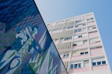A tall building with a modern architectural style, featuring a pastel-colored facade with multiple windows. Adjacent to the building is a large, colorful mural comprised of geometric shapes and abstract figures in shades of blue, green, and purple.