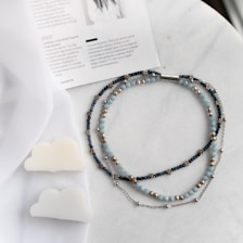 A collection of beaded necklaces with blue, silver, and gold beads is arranged on a white surface next to a magazine. Two cloud-shaped candles are also present, with a soft fabric draped partially over them.