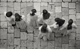 A group of seven children wearing light-colored clothing stand on a stone-tiled pavement. Their backs are turned as they appear to be lined up or walking in unison, with some of the children having long hair or floral accessories.