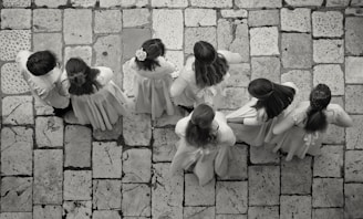A group of seven children wearing light-colored clothing stand on a stone-tiled pavement. Their backs are turned as they appear to be lined up or walking in unison, with some of the children having long hair or floral accessories.
