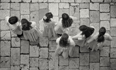 A group of seven children wearing light-colored clothing stand on a stone-tiled pavement. Their backs are turned as they appear to be lined up or walking in unison, with some of the children having long hair or floral accessories.