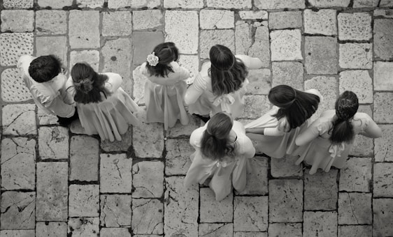 A group of seven children wearing light-colored clothing stand on a stone-tiled pavement. Their backs are turned as they appear to be lined up or walking in unison, with some of the children having long hair or floral accessories.