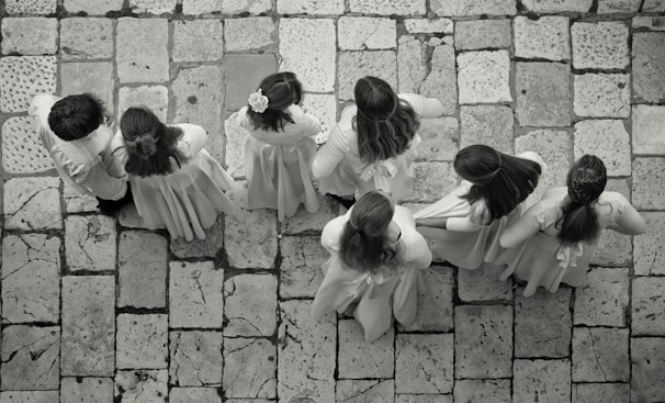 A group of seven children wearing light-colored clothing stand on a stone-tiled pavement. Their backs are turned as they appear to be lined up or walking in unison, with some of the children having long hair or floral accessories.