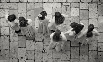 A group of seven children wearing light-colored clothing stand on a stone-tiled pavement. Their backs are turned as they appear to be lined up or walking in unison, with some of the children having long hair or floral accessories.