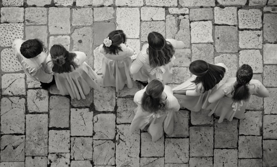 A group of seven children wearing light-colored clothing stand on a stone-tiled pavement. Their backs are turned as they appear to be lined up or walking in unison, with some of the children having long hair or floral accessories.