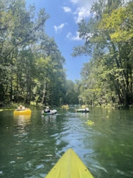 A serene river scene with several people relaxing on inflatable rafts and tubes, surrounded by lush greenery and tall trees under a bright blue sky with scattered clouds. The foreground features the tip of a yellow kayak, suggesting a perspective from someone paddling.