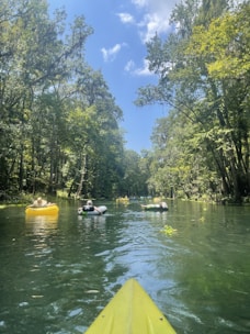A serene river scene with several people relaxing on inflatable rafts and tubes, surrounded by lush greenery and tall trees under a bright blue sky with scattered clouds. The foreground features the tip of a yellow kayak, suggesting a perspective from someone paddling.