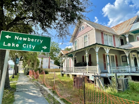 A historic, two-story house with an ornate wooden porch and a corrugated metal roof sits along a sidewalk. There is a green road sign nearby indicating directions to Newberry and Lake City. The area is surrounded by trees and a few other buildings, while an orange safety net separates the sidewalk from the grass.