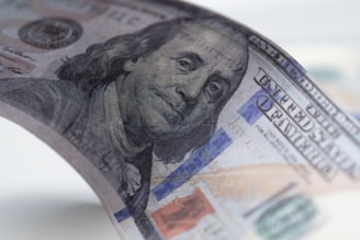 A close-up view of a United States hundred-dollar bill, featuring a partial image of Benjamin Franklin's face. The bill is slightly curved, with visible details like the blue security ribbon and intricate background patterns.