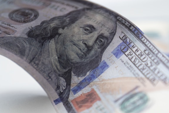 A close-up view of a United States hundred-dollar bill, featuring a partial image of Benjamin Franklin's face. The bill is slightly curved, with visible details like the blue security ribbon and intricate background patterns.